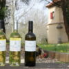 trois bouteilles de vin blanc posées sur un banc en bois avec un pigeonnier périgourdin en arrière plan et un coq qui picore dans l'herbe